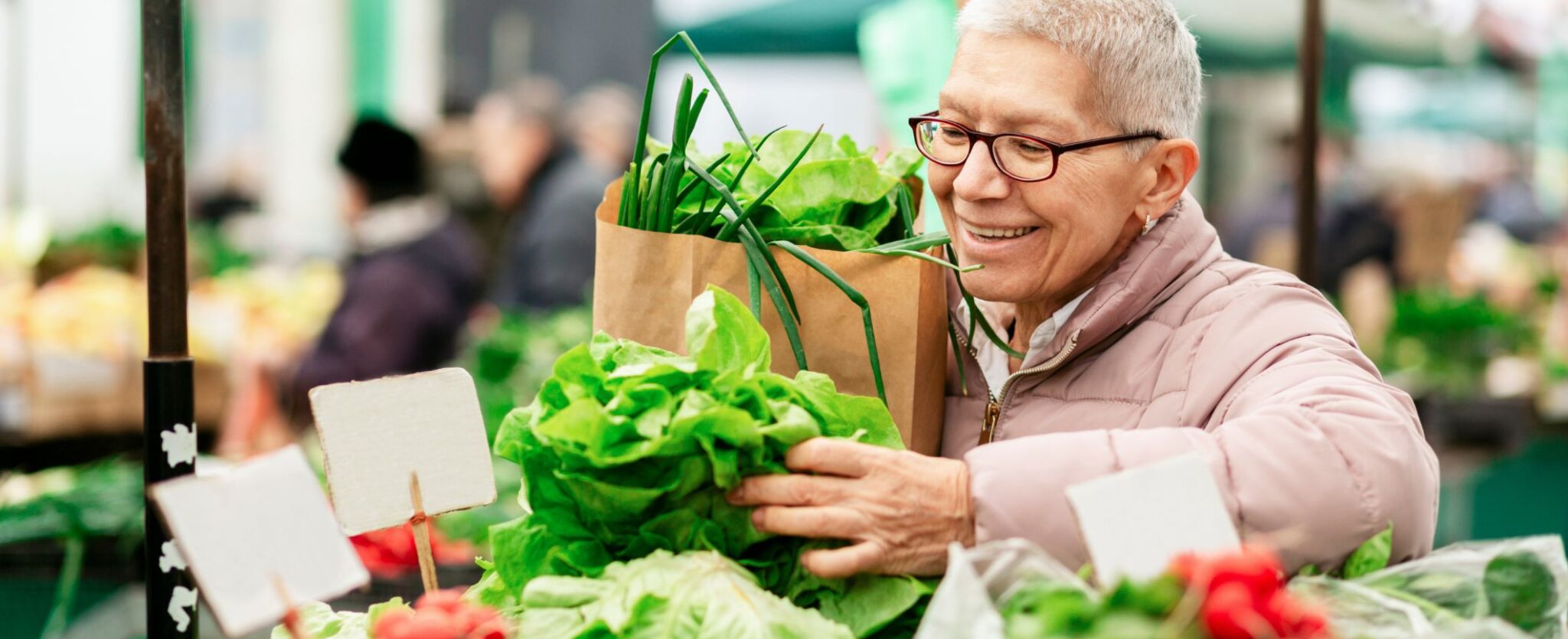 An older woman smiling as she holds a bunch of fresh lettuce and a brown paper bag filled with produce at an outdoor market.